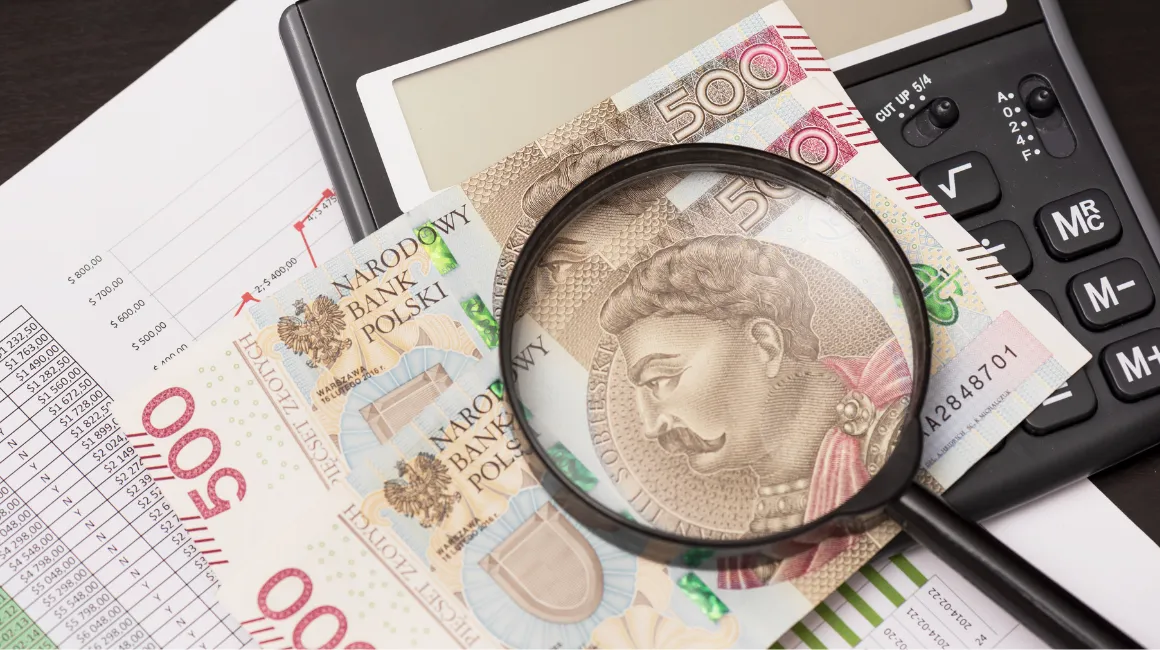 Zbliżenie na banknoty 500 zł z lupą powiększającą fragment portretu, leżące obok kalkulatora i arkuszy z wykresami finansowymi