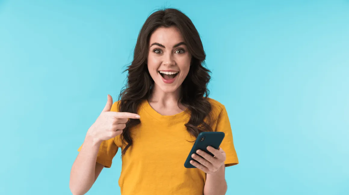 Uśmiechnięta młoda kobieta w żółtej koszulce, trzymająca smartfon i wskazująca na niego z entuzjazmem na tle błękitnej ściany.